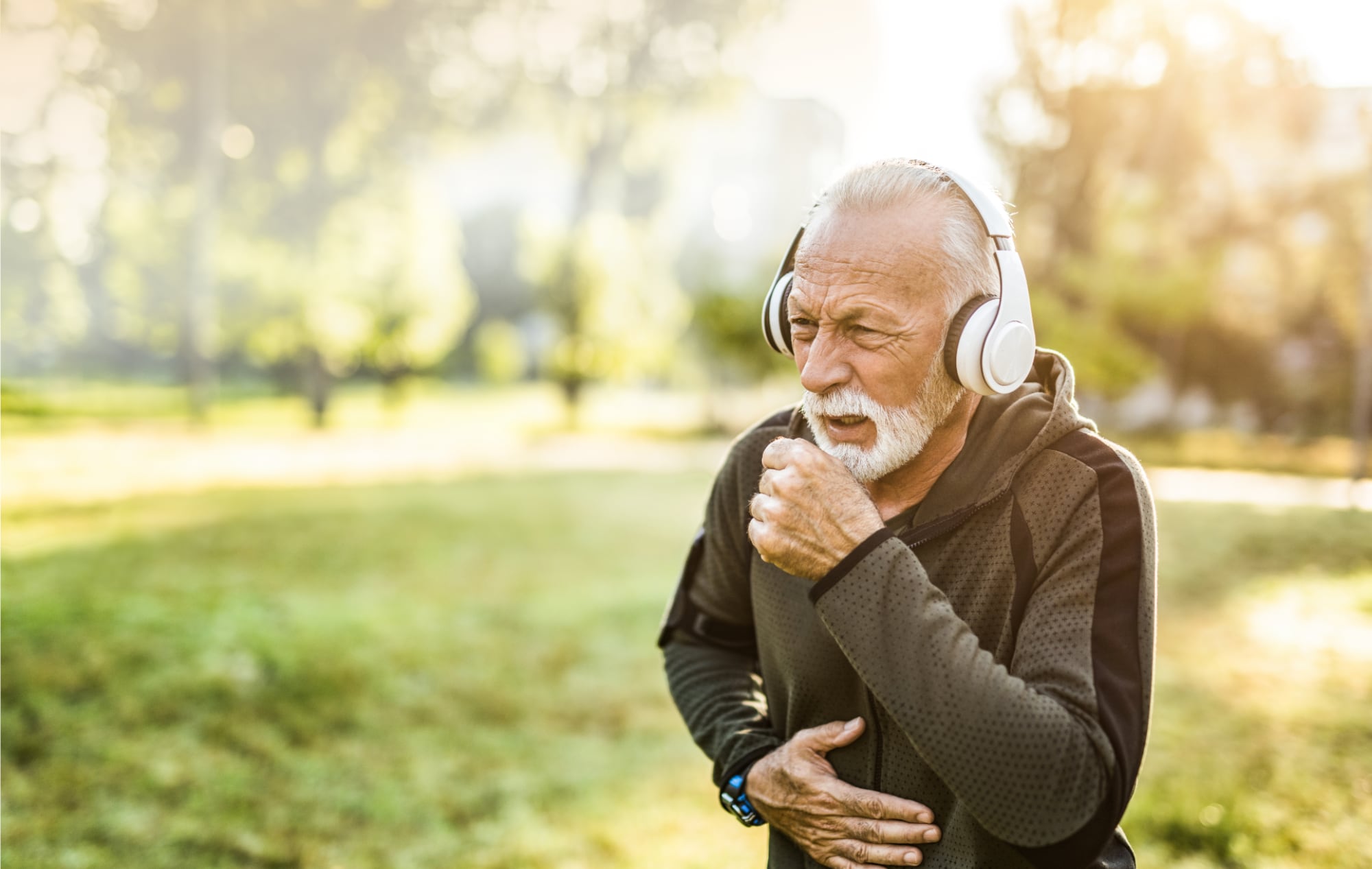 Senior man with headphones while jogging in a park coughs into his hand.